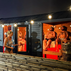 Outdoor evening scene of a modern sauna facility with string lights overhead. The structure features dark exterior cladding with three illuminated sauna rooms with orange-tinted warmth visible through glass doors. People in swimwear are visible - one woman standing outside conversing with someone in a sauna, and two men sitting together in another sauna room. The facility has wooden decking and a curved roof, creating an inviting wellness space.