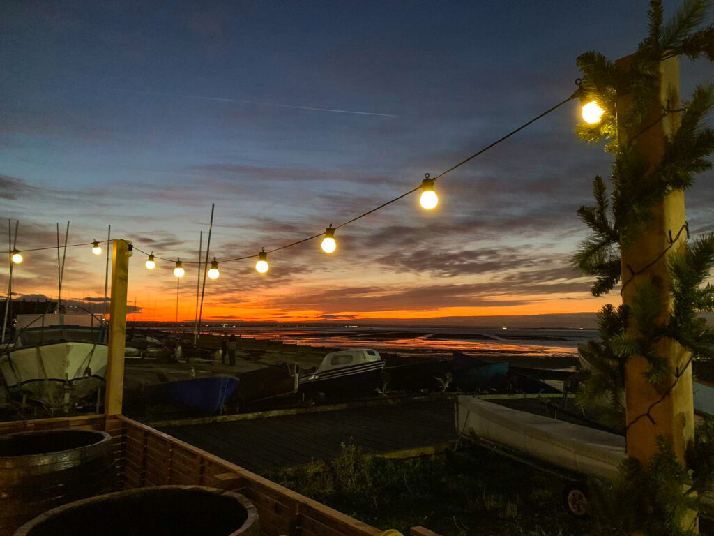 Sea Scrub Sauna Whitstable Harbour location at sunset during Autumn Equinox celebration with string lights, cold plunge barrels, boats and dramatic orange sky over Kent coast