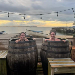 Two women cold plunging in whisky barrels at Whitstable Harbour at sunset, Sea Scrub Sauna Kent